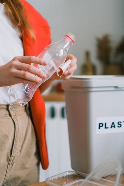 A person wearing a red cardigan and beige trousers is holding a clear plastic bottle near the top, preparing to dispose of it into a large grey recycling bin labeled 'PLASTIC'. The environment appears