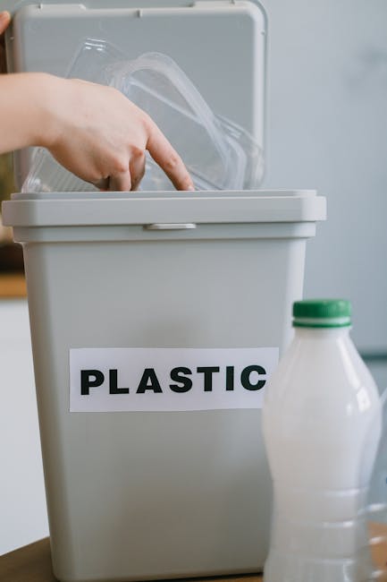 A person wearing a red cardigan and beige trousers is holding a clear plastic bottle near the top, preparing to dispose of it into a large grey recycling bin labeled 'PLASTIC'. The environment appears