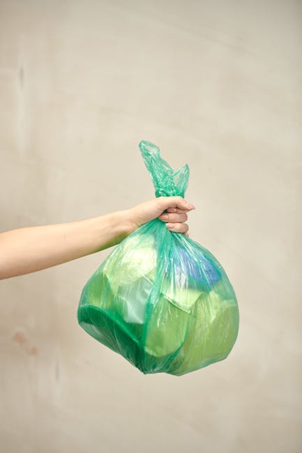 A person's hand holding a green and blue plastic shopping bag filled with household items, likely related to home relocation or packing. The bag appears to contain soft materials such as clothing or fabric, and is being held in front of a plain, light-colored background. This image illustrates the process of packing or decluttering in preparation for a move, aligning with furniture disposal or removal services offered by Putney Removals as part of house removals or moving logistics. The bag's crumpled appearance and the way it is gripped suggests careful handling during packing or transportation, emblematic of packing and loading processes involved in furniture transport or home relocations.