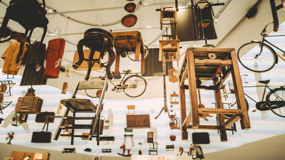 An indoor exhibit space features an array of vintage and modern household items suspended from the ceiling and mounted on the walls. Visible objects include wooden chairs, small tables, bicycles, suitcases, and various boxes, some wrapped in plastic or fabric. The items are arranged at different heights, with some on the floor, others hanging from ceiling hooks, and a few resting on shelves. A bicycle is positioned upright on the right side, while a wooden chair and a small table are prominently displayed in the foreground, suggesting a focus on furniture collection. The environment is well-lit with natural light coming through windows, casting soft shadows and highlighting the textures of wood and metal objects. This eclectic display reflects a carefully curated collection of items that would be typical in a furniture or household goods store, relevant for home relocation and furniture transport services. Putney Removals specializes in managing such bulky-item collections as part of their house removal and packing services, ensuring safe and efficient transportation during relocation.