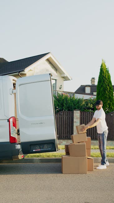A man wearing a black cap, white T-shirt, and jeans sitting on the pavement beside an open van loaded with various cardboard boxes of different sizes, some sealed with packing tape and others partiall