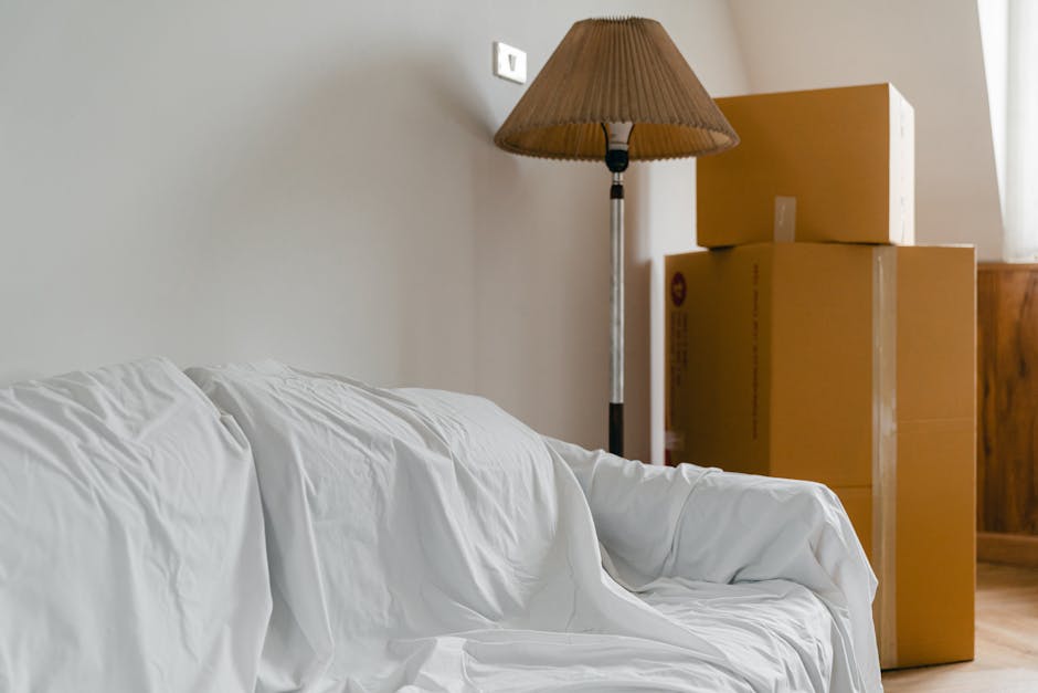 The image shows a living room corner with a white sofa covered by a crumpled white sheet, situated in front of a plain light-colored wall. To the right of the sofa, there are three large cardboard boxes stacked on top of each other, with the top box slightly smaller than the two below. A tall floor lamp with a brown fabric lampshade stands behind the boxes, casting soft light on the scene. The boxes are packed with packing materials such as plastic wrap and are positioned near a wooden piece of furniture, possibly a sideboard or cabinet, suggesting a home relocation process. The setting indicates an active packing and moving environment, with furniture and belongings prepared for transport, which aligns with the context of house removals and the focus on avoiding hidden fees in Putney removals. The overall scene reflects the logistical aspect of furniture transport and packing for a home move.