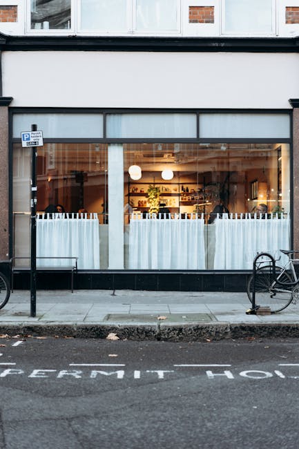 A large glass storefront window of a café or restaurant located on the ground floor of a building, with white curtains hanging inside and warm interior lighting visible through the glass. The pavement in front of the shop features a partially visible parking area with the words 'PERMIT HOLDER' painted on the asphalt. Two bicycles are secured to metal bike racks on either side of the entrance. The building's exterior includes a brick wall above the window and modern framing around the glass. Inside, shelves with various items are visible, and the scene suggests an urban setting suitable for home relocation planning, such as business removals or furniture transport tasks, with natural daylight illuminating the exterior. This image is relevant to Putney Removals' services related to moving logistics and street permits, highlighting the importance of careful planning when relocating home or business belongings within a street setting.