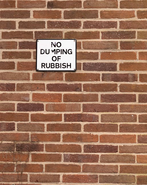 A rectangular white metal sign with black uppercase text that reads 'NO DUMPING OF RUBBISH' is mounted on a red brick wall with a traditional running bond pattern. The bricks vary in shades of red and brown, and the mortar joints are light grey. The sign is centered horizontally on the wall, approximately at eye level, and appears to be securely attached. The wall's surface is clean with no visible graffiti or damage. This setting could be relevant to home relocation or moving services, as it depicts a typical external building feature, possibly in a residential area, where parking or access regulations might be important for furniture transport and loading logistics, which are part of the services provided by Putney Removals.