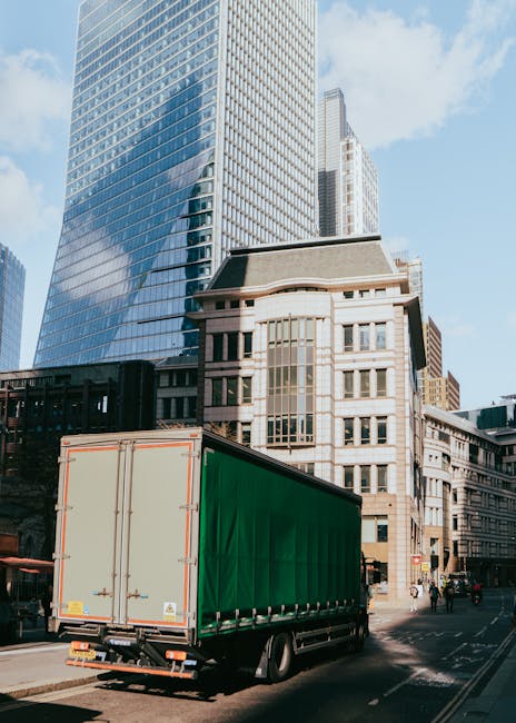 A large commercial delivery truck with a green cargo area is parked on a city street in Putney, surrounded by tall modern office buildings and older historic structures. The truck is positioned at the curb, partially on the road, with the rear doors closed. The surrounding environment features clear blue skies and a few pedestrians and cyclists on the pavement. This scene depicts a home relocation or furniture transport process as part of a moving service, with the vehicle ready for loading or unloading furniture, boxes, or packing materials typically used during packing and moving activities. The cityscape backdrop emphasizes the urban context of house removals and logistics, with the presence of tall glass buildings and older brick and stone facades illustrating an area where professional removals, such as those provided by Putney Removals, operate efficiently.