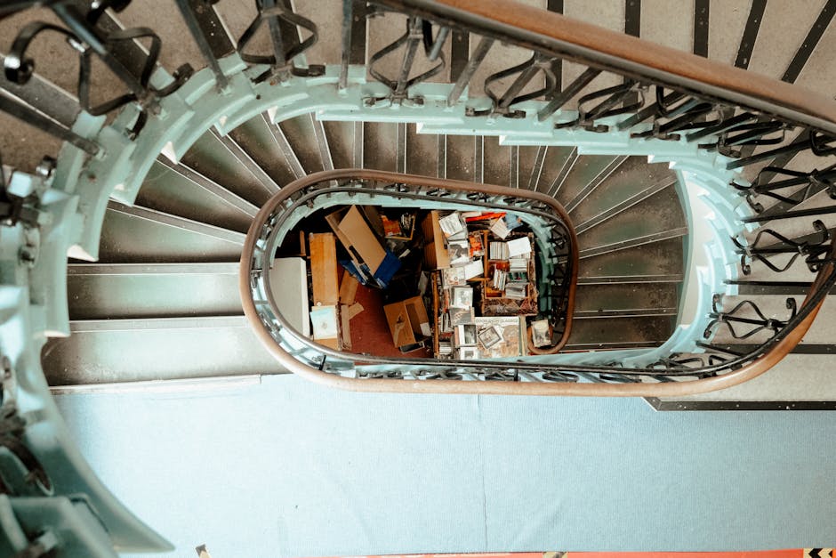 View from the top of a spiral staircase looking downward into the stairwell, showcasing a pile of boxes, cardboard cartons, and packing materials arranged on the floor below. The staircase features a metal railing with black wrought iron balusters and a polished wooden handrail. The steps are made of dark-colored material, possibly wood or painted metal, with visible treads. Natural light illuminates the scene, highlighting the clutter of various-sized boxes, some open with contents visible, indicative of a home relocation or packing process. The surrounding walls are light-colored, and part of the stairway’s blue carpeted surface is visible at the top. This setup presents a typical staircase layout that might pose challenges for furniture transport during a house move, as illustrated by Putney Removals on their webpage about staircase, parking, and lift challenges for Putney moves.