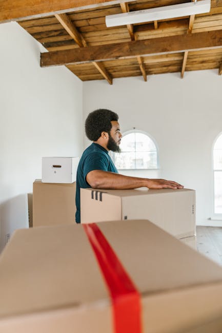 A man with a dark beard and curly hair, wearing a teal shirt, stands inside a spacious room with white walls and a wooden ceiling with exposed beams. He is positioned behind several stacked cardboard moving boxes, some sealed with red packing tape, preparing for a home relocation. The room has large, arched windows allowing natural light to enter, and the man appears to be in the process of organising or preparing items for packing and moving with the assistance of Putney Removals. The boxes are arranged on the floor, indicating the initial stages of a furniture transport and packing process, typical of professional removals services in the SW15 area.