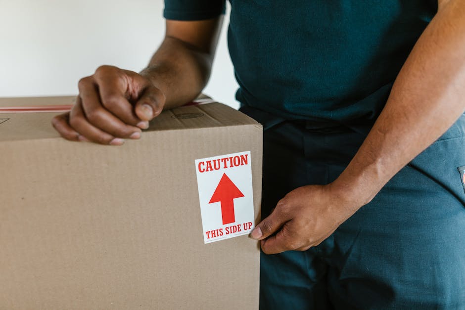 Close-up of a person wearing a dark green T-shirt and dark trousers, preparing a cardboard moving box by securing a red and white caution label indicating the top side with an upward arrow, during a home relocation process. The person’s hands are visible holding the box firmly, with the background partially showing a doorway and a light-colored interior wall, suggesting an indoor environment within a property being prepared for moving. The scene highlights packing and loading activities typical of furniture transport and professional removals services by Putney Removals in the context of a house move, emphasizing careful handling and proper labeling of boxes for safe transport.