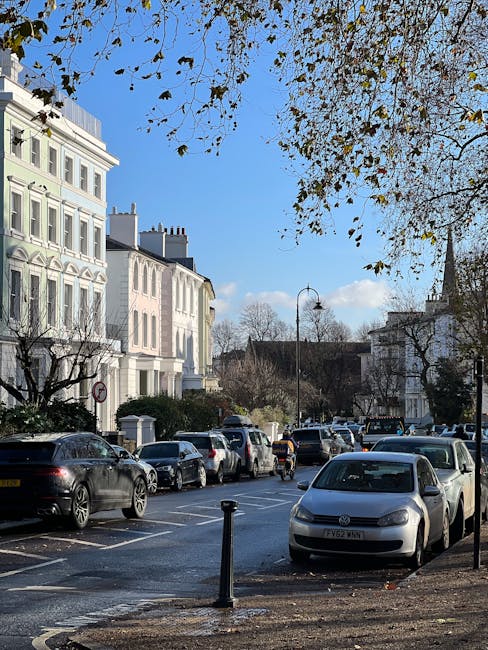 A street scene on Upper Richmond Road in Putney showing a row of white residential buildings with decorative facades and large windows, parked cars lining both sides of the road including small hatchbacks and larger vehicles, some with visible roof racks and open doors. There is a tree in the foreground with branches extending across the top of the image, some leaves still attached while others have fallen. A metal bollard is positioned at the edge of the pavement, and several cardboard boxes, wrapped furniture items, and protective padding are visible near the parked cars, indicating preparations for a home relocation. The sky is bright with scattered clouds, and a lamppost stands along the street, reflecting typical London residential neighbourhood features. The scene captures the environment during daylight hours, with a focus on vehicle parking and street frontage suitable for loading of furniture and moving equipment, as coordinated by Putney Removals for house removals and logistics.