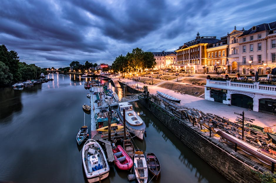 A nighttime scene of a marina along the Upper Richmond Road in Putney showing boats docked on a calm river with reflections of the vessels on the water. On the riverside promenade, there are several piles of wooden pallets and stacked ladders, with some plastic and cardboard packing materials nearby, indicating recent packing activities related to house removals. A row of well-lit, multi-storey residential buildings with elegant architecture lines the street, with outdoor street lamps casting warm light. The pavement area is mostly clear, while the boats are secured along the quay, suggesting a pause in the loading or unloading process associated with home relocation or furniture transport. The cloudy sky overhead adds a moody atmosphere, and occasional boat lights flicker on the tranquil water surface, emphasizing the quiet yet active environment connected to house removals near the river, as managed by Putney Removals.