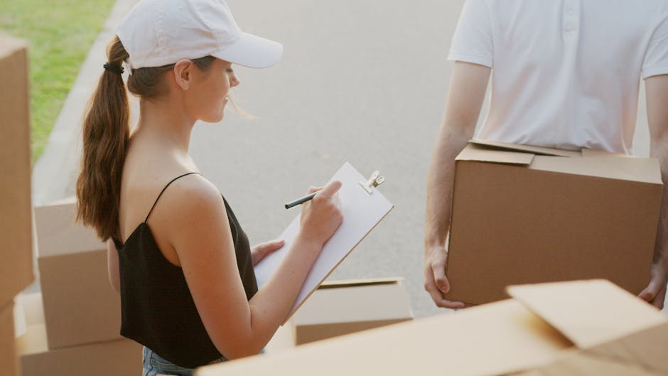 A young woman with a white cap and black spaghetti strap top is holding a clipboard and pen, recording information during a house removal process. She is standing outdoors near a paved area with several carbon cardboard boxes, some open and others sealed with packing tape, placed on a flat surface. To her right, a person in a white T-shirt is holding a medium-sized cardboard box, assisting in the packing and moving activities. The scene indicates an organized packing phase of a home relocation, with light natural daylight illuminating the area, and the background showing a blurred outdoor environment. The presence of packing materials and the ongoing handling of boxes suggest a furniture transport and household items packing process, typical of professional removals services such as those offered by Putney Removals. This setting reflects the logistical aspects of home relocation, including packing, loading, and transport preparation.